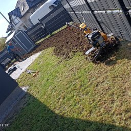 Klaudia Kajtarek - Przygotowanie terenu pod trawnik: glebogryzarka spalinowa w akcji, mężczyzna z grabiami wyrównuje ziemię, widoczny fragment metalowego ogrodzenia panelowego i budynek mieszkalny w tle.