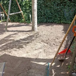 Przygotowany teren pod trawnik z widocznym drzewem pośrodku, narzędziami ogrodniczymi (grabie, łopata) i konstrukcją huśtawki w tle, otoczony zielonym bluszczem.