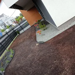 Świeżo założony trawnik z brązową korą, obok nowoczesnego domu z szarą elewacją, czarnym ogrodzeniem i rabatami z kamykami oraz zielenią.