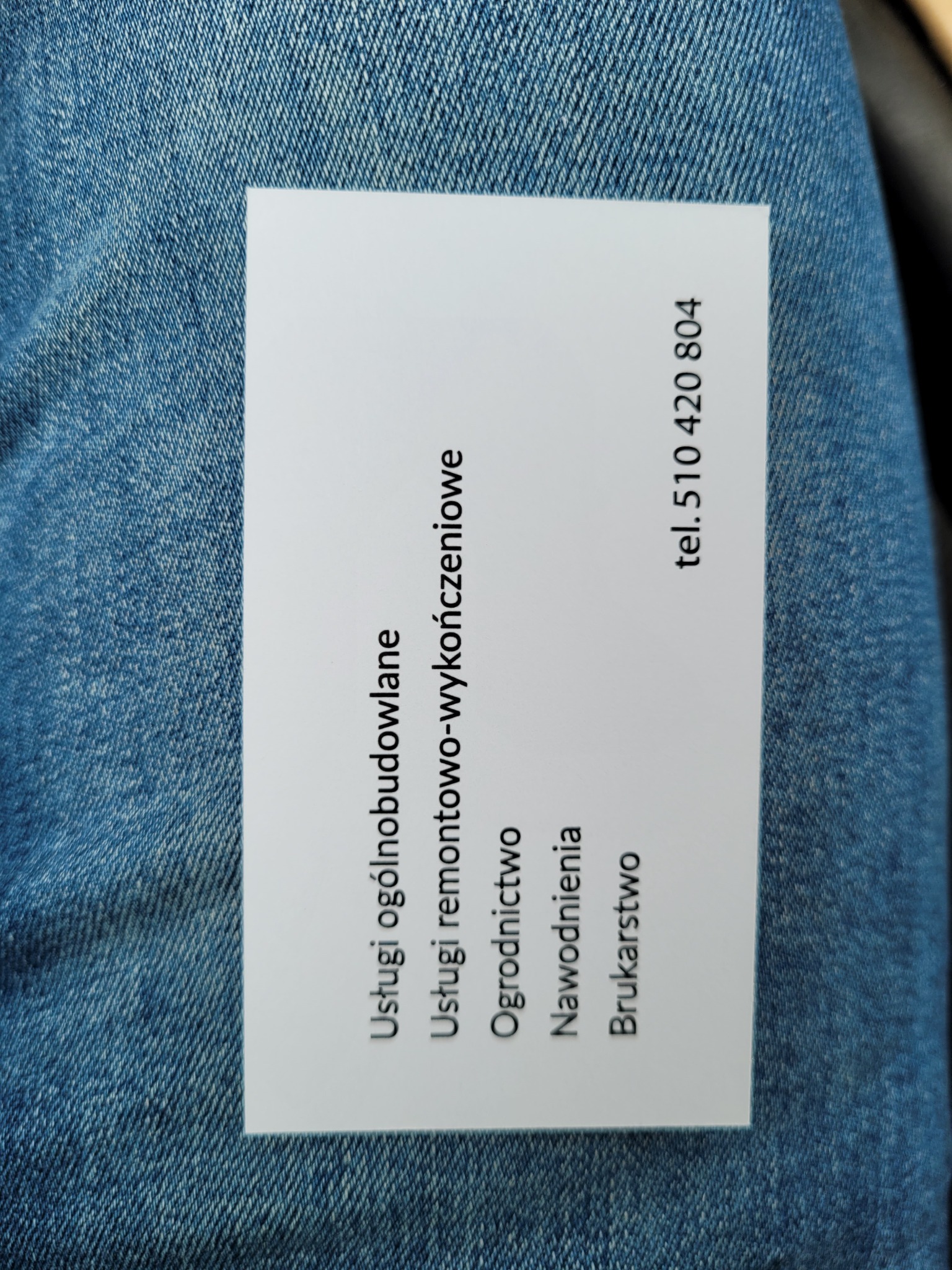 Wizytówka firmy z usługami ogólnobudowlanymi, remontowo-wykończeniowymi, ogrodnictwem, nawodnieniami i brukarstwem, leżąca na dżinsach.
