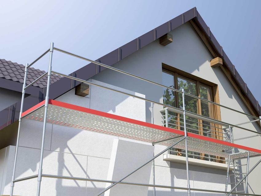 Dom jednorodzinny w trakcie szpachlowania elewacji, widoczne rusztowanie z czerwonymi elementami, okno z drewnianą ramą i fragment dachu z brązowej dachówki.