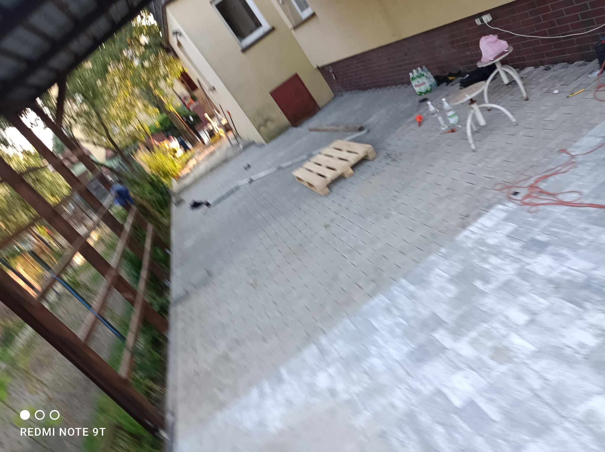 Niedokończona nawierzchnia z szarej kostki brukowej przed budynkiem, widoczna paleta, narzędzia i akcesoria budowlane, perspektywa z góry.