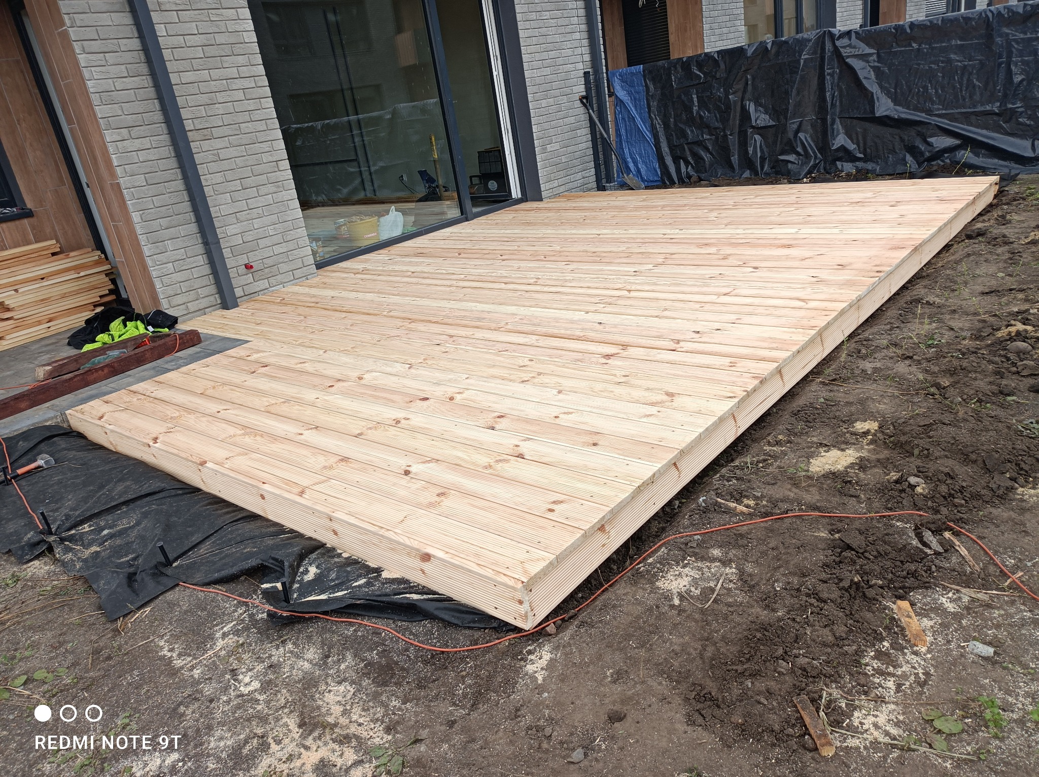 Drewniany taras w trakcie budowy, widoczne deski tworzące powierzchnię, czarna folia pod spodem, narzędzia i materiały budowlane obok, ziemia wokół.