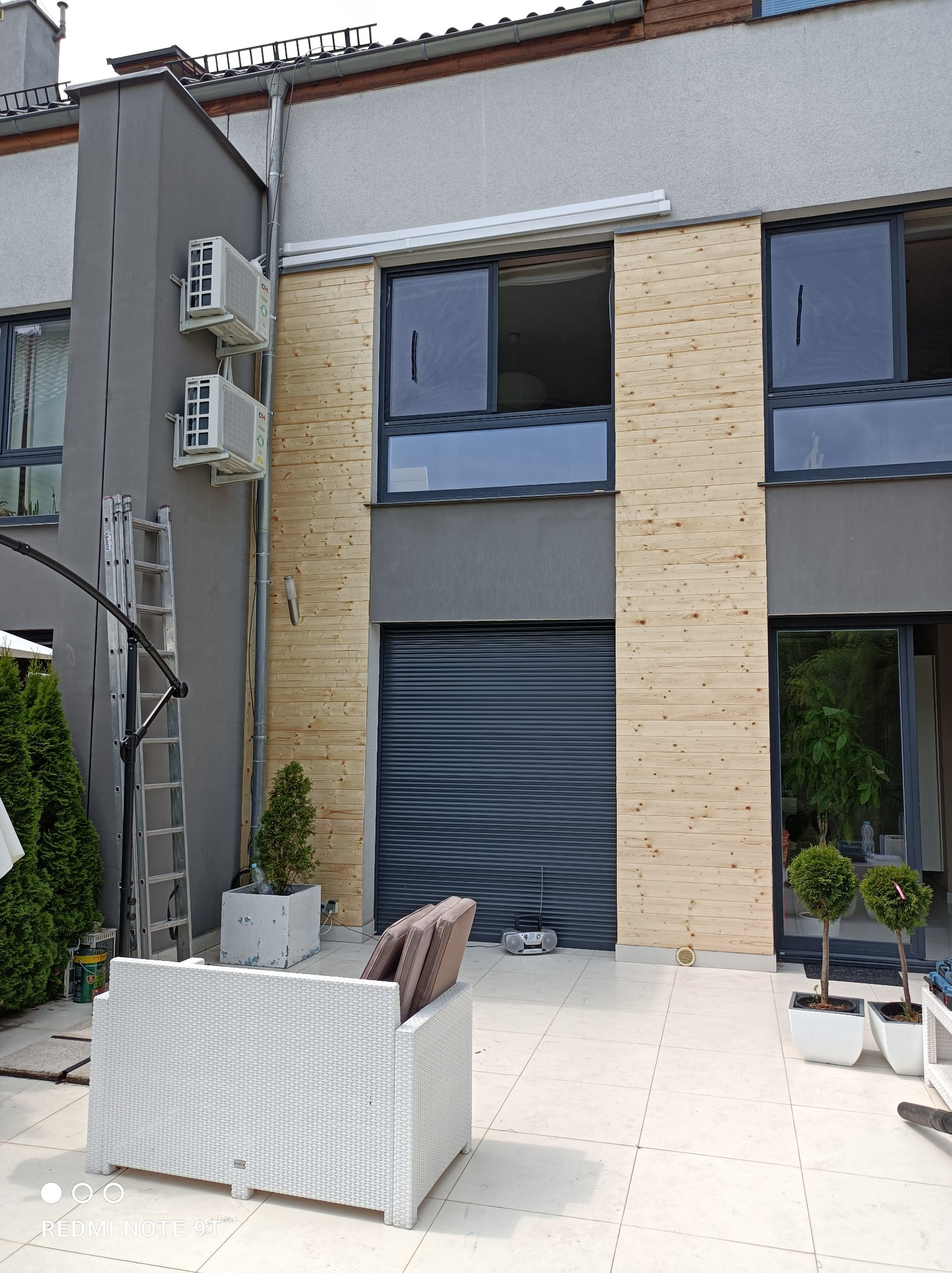Elewacja budynku z drewnianą okładziną, widoczne okna, brama garażowa, klimatyzatory na szarej ścianie, drabina oparta o ścianę, białe meble ogrodowe na jasnym tarasie.