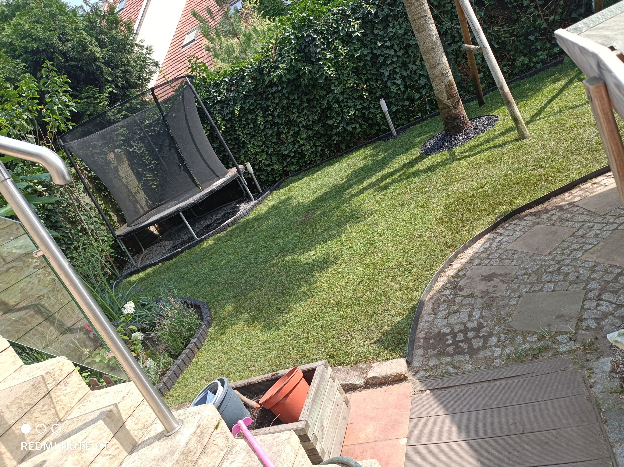 Świeżo założony trawnik z trampoliną w tle, obrzeża z kostki, donice z kwiatami i schody z balustradą.