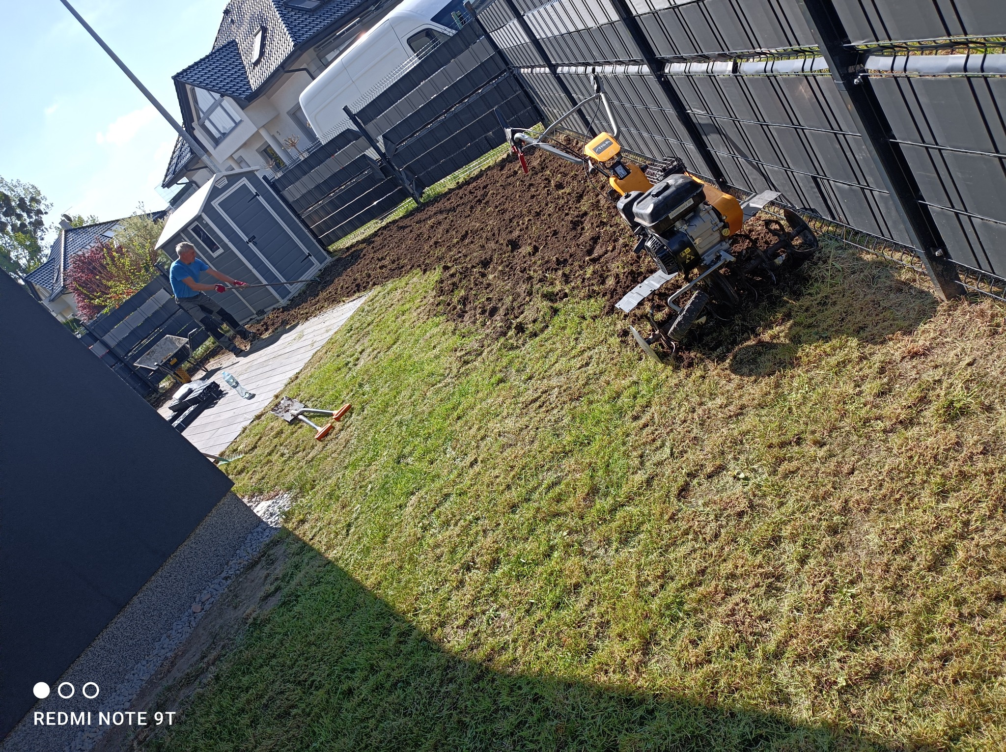 Przygotowanie terenu pod trawnik: glebogryzarka spalinowa w akcji, mężczyzna z grabiami wyrównuje ziemię, widoczny fragment metalowego ogrodzenia panelowego i budynek mieszkalny w tle.