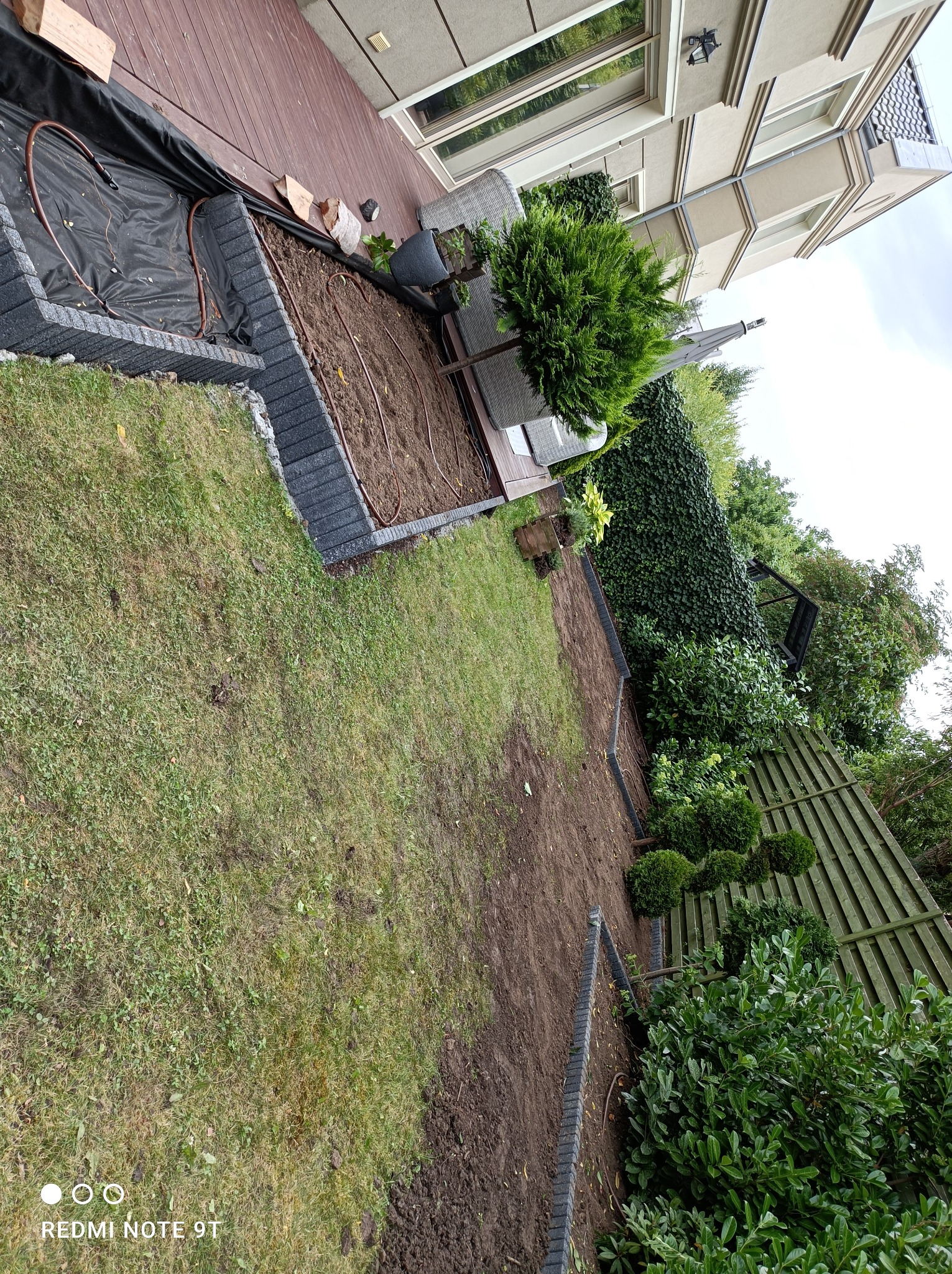 Przygotowanie terenu pod trawnik z nawadnianiem, widoczne obrzeża z kostki, rozłożona instalacja nawadniająca i czarna folia, trawa w tle, dom z jasną elewacją w oddali.