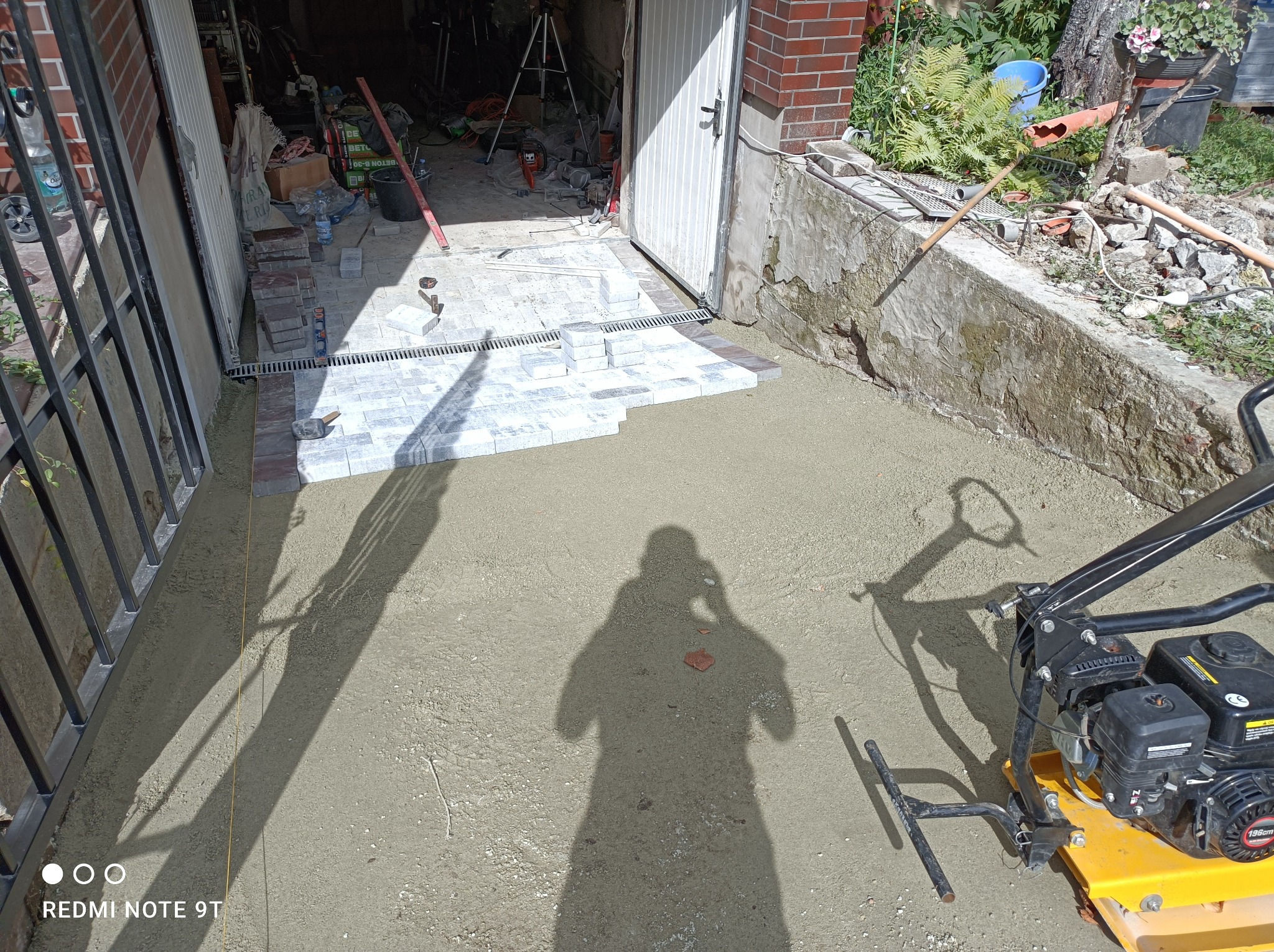 Fragment podjazdu z ułożoną częścią jasnoszarej kostki brukowej, widoczny żółty zagęszczarką, przygotowane podłoże, brama garażowa w tle, cień osoby robiącej zdjęcie.
