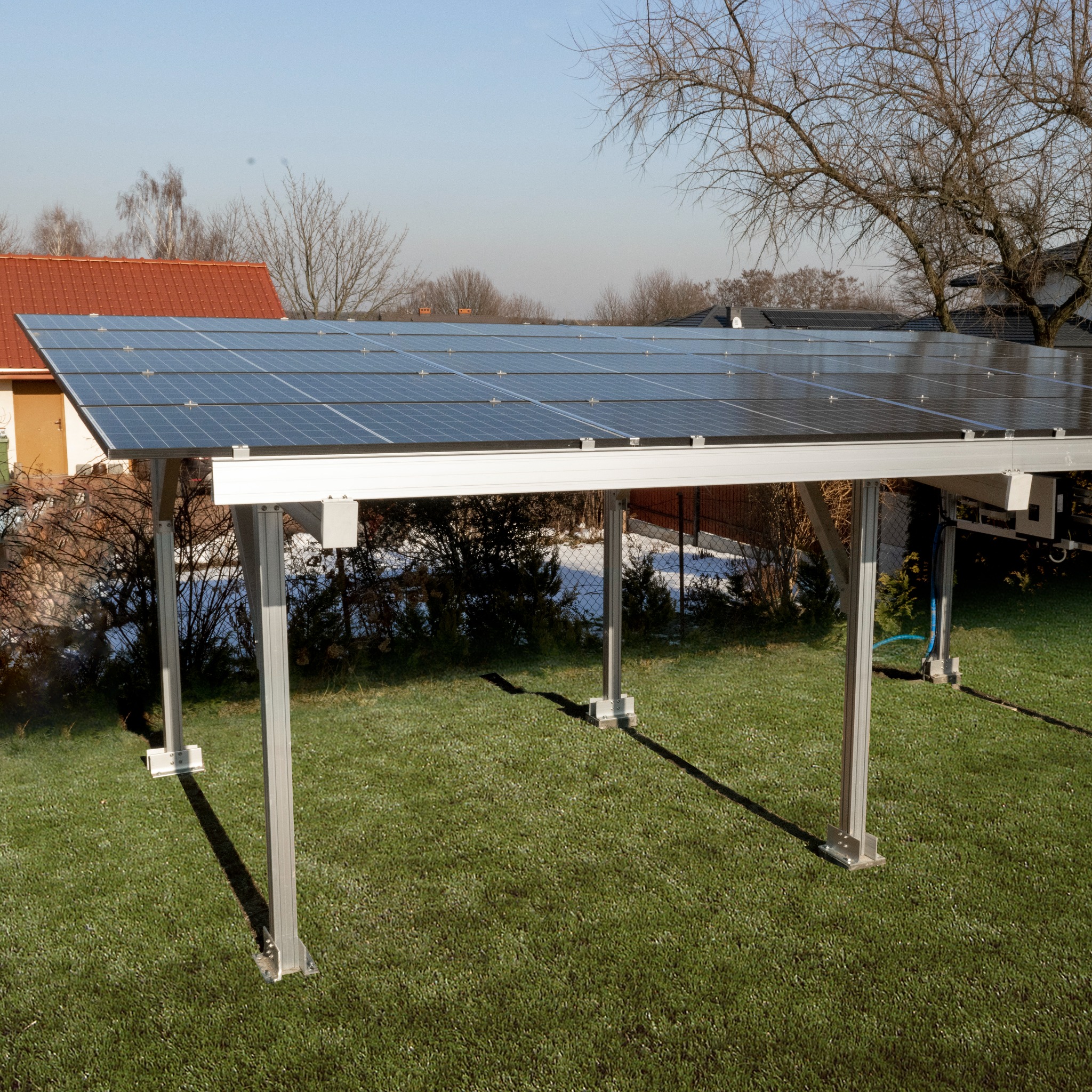 Nowoczesna wiata fotowoltaiczna na aluminiowej konstrukcji, z panelami słonecznymi, zainstalowana na trawniku przed domem.