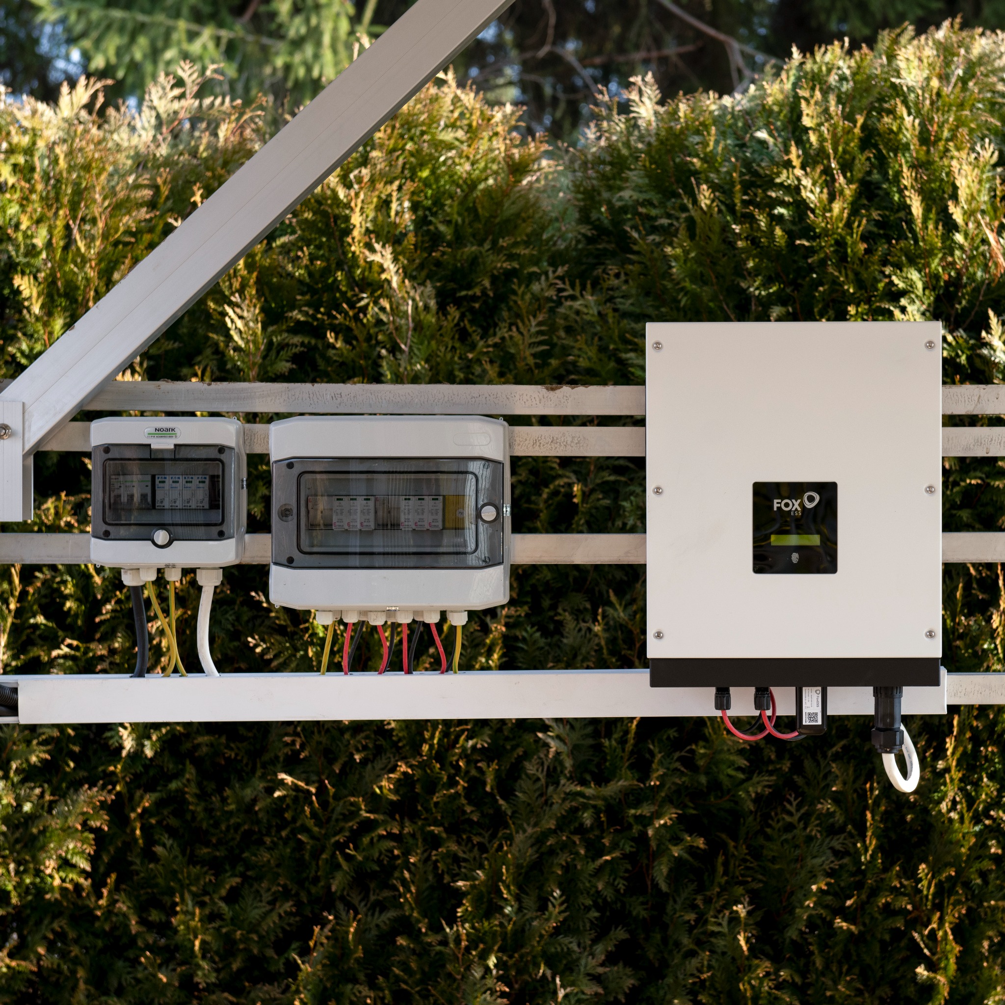 Instalacja fotowoltaiczna: skrzynki zabezpieczeń i falownik Fox ESS zamontowane na konstrukcji na tle zielonych tuj.