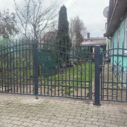 Nowoczesna, grafitowa brama wjazdowa z furtką, zdobiona delikatnymi ornamentami, na tle zieleni ogrodu i jasnoniebieskiego domu z drewnianą elewacją, ułożona na kostce brukowej.