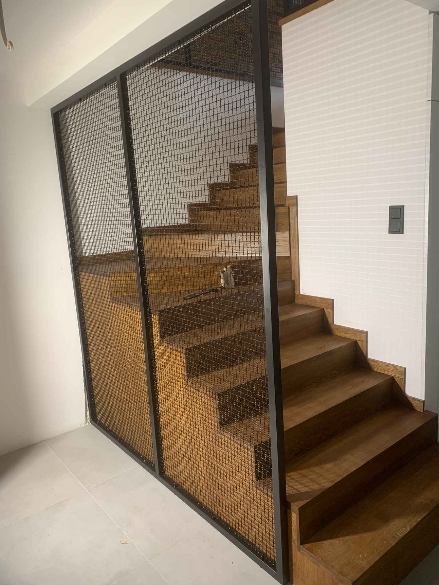 Drewniane schody z metalową siatką ochronną, widok z dołu, nowoczesny design, białe ściany i płytki podłogowe.