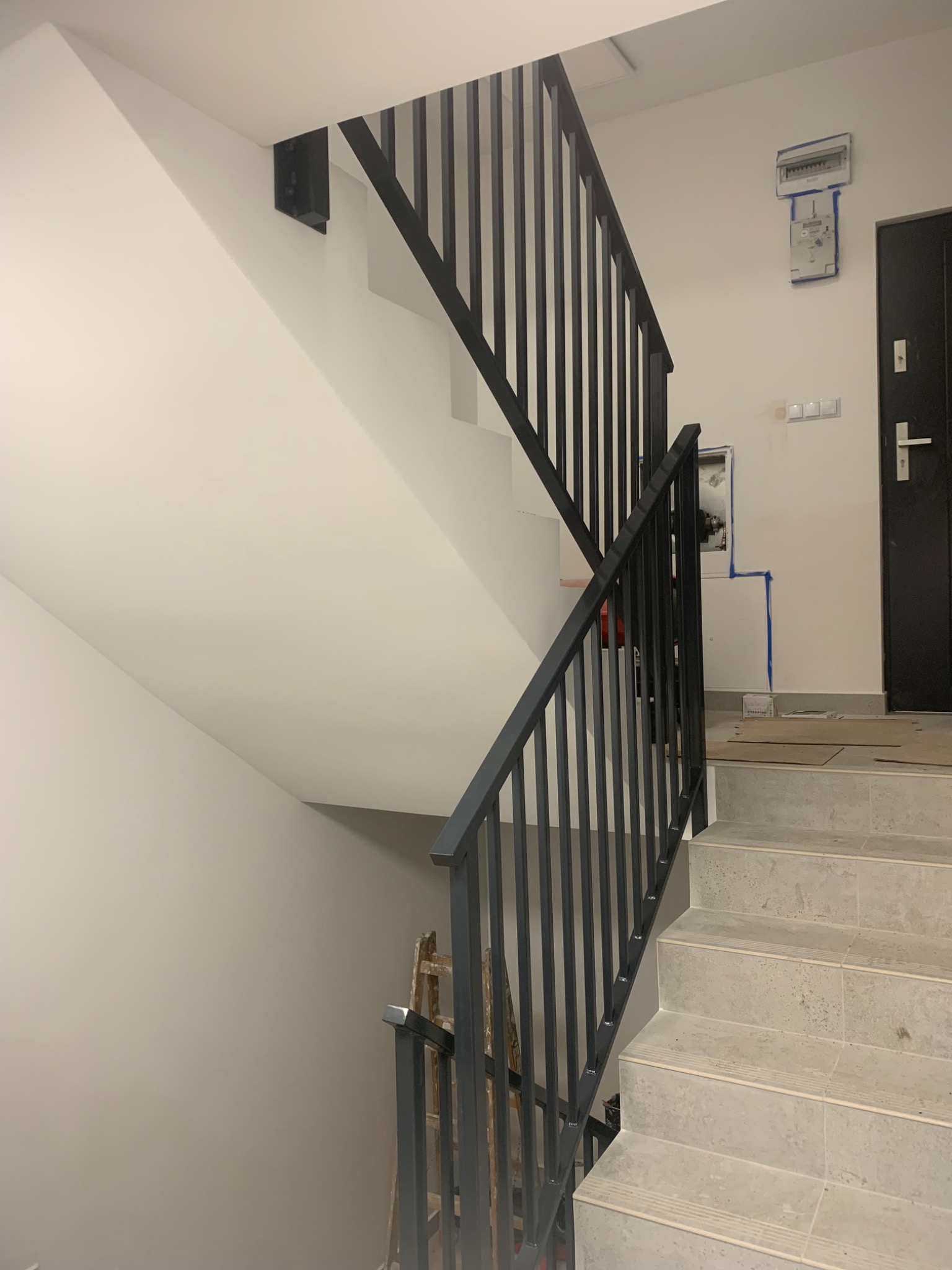 Nowoczesne schody z szarymi płytkami i czarną metalową balustradą, widoczne zabezpieczenia na ścianie i drabina w tle.