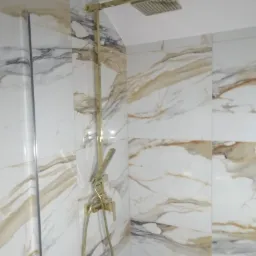 Błyszcząca, złota armatura prysznicowa z deszczownicą na ścianie wyłożonej białymi płytkami imitującymi marmur z brązowymi żyłami.