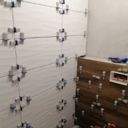 Białe, faliste płytki ścienne zamocowane krzyżykami dystansowymi w trakcie montażu, z widocznymi instalacjami hydraulicznymi i fragmentem drewnopodobnej okładziny na sąsiedniej ścianie.