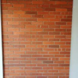 Ściana z czerwonej cegły, ułożona w regularny wzór, z widocznym białym fragmentem ściany po lewej stronie kadru.