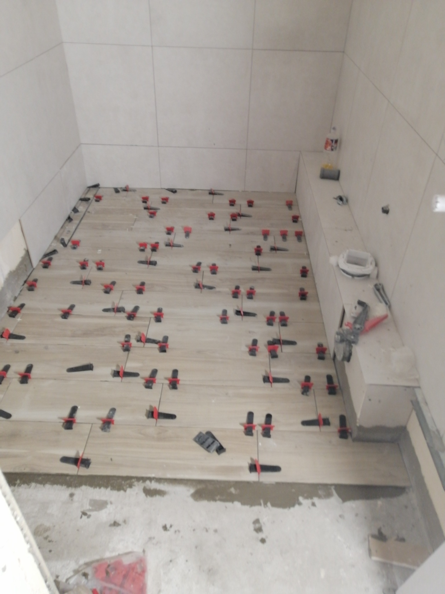 Podczas prac w łazience: ułożone jasne płytki podłogowe z systemem poziomowania, widoczne narzędzia i akcesoria na półce.