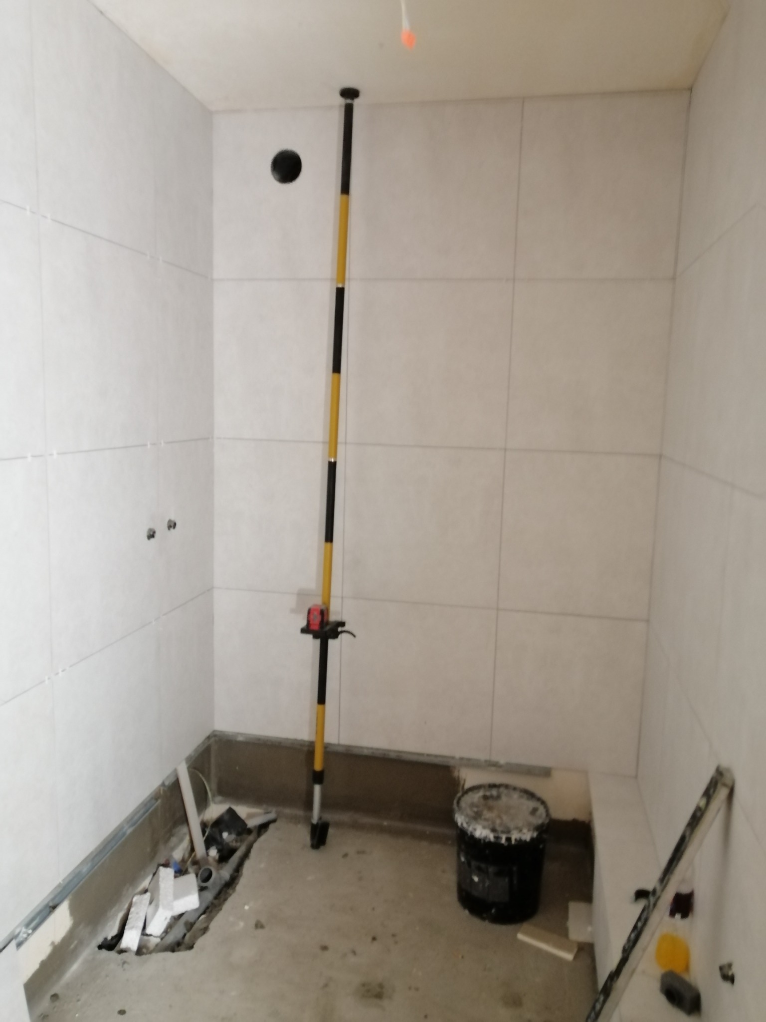 Wnętrze łazienki w trakcie prac, z jasnymi, kwadratowymi płytkami na ścianach, poziomica laserowa ustawiona na podłodze, widoczne przygotowane miejsce na brodzik oraz wiadro z zaprawą.