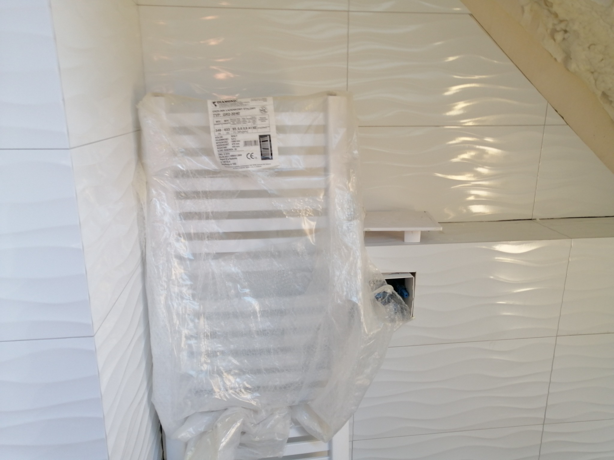 Biały grzejnik łazienkowy, jeszcze w folii ochronnej, zamontowany na ścianie z białych, falowanych płytek ceramicznych w łazience ze skośnym sufitem.