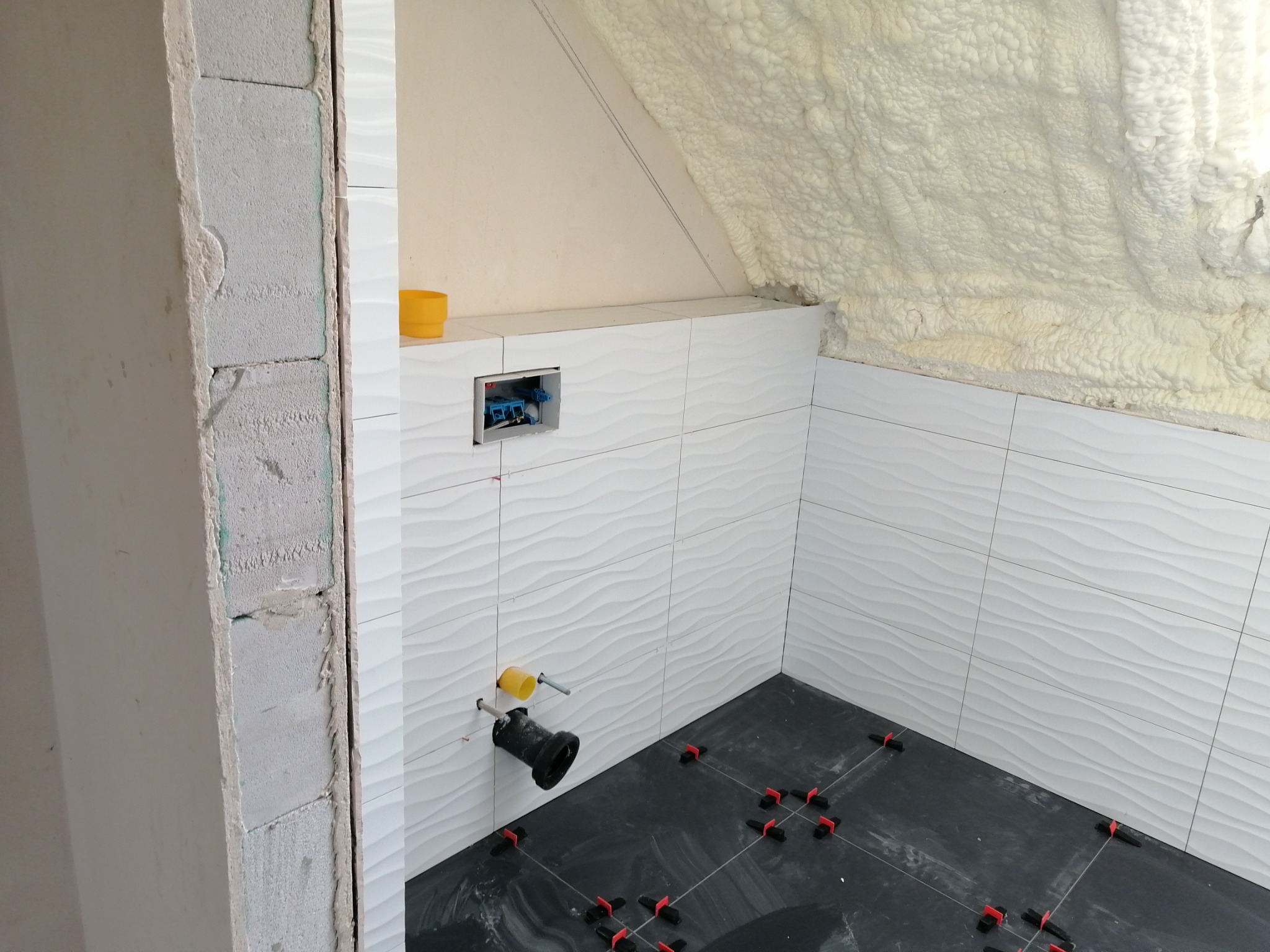 Białe, faliste płytki ścienne w łazience na poddaszu, czarne płytki podłogowe z czerwonymi krzyżykami dystansowymi, widoczna instalacja podtynkowego WC, ocieplenie pianką na skośnym suficie.