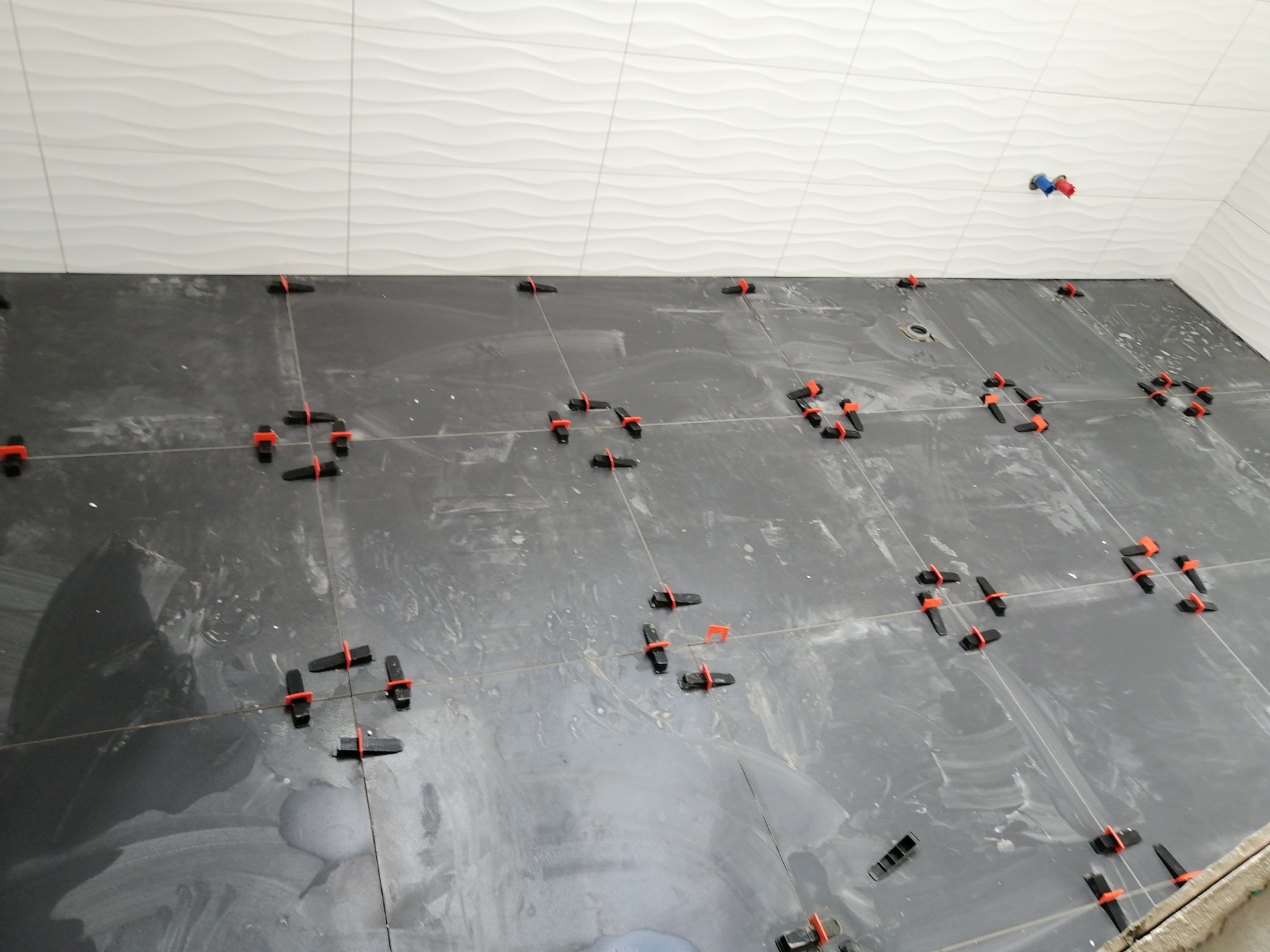 Ciemnoszare płytki podłogowe w trakcie układania, z widocznymi krzyżykami dystansowymi, na ścianie białe płytki z delikatnym wzorem fal.