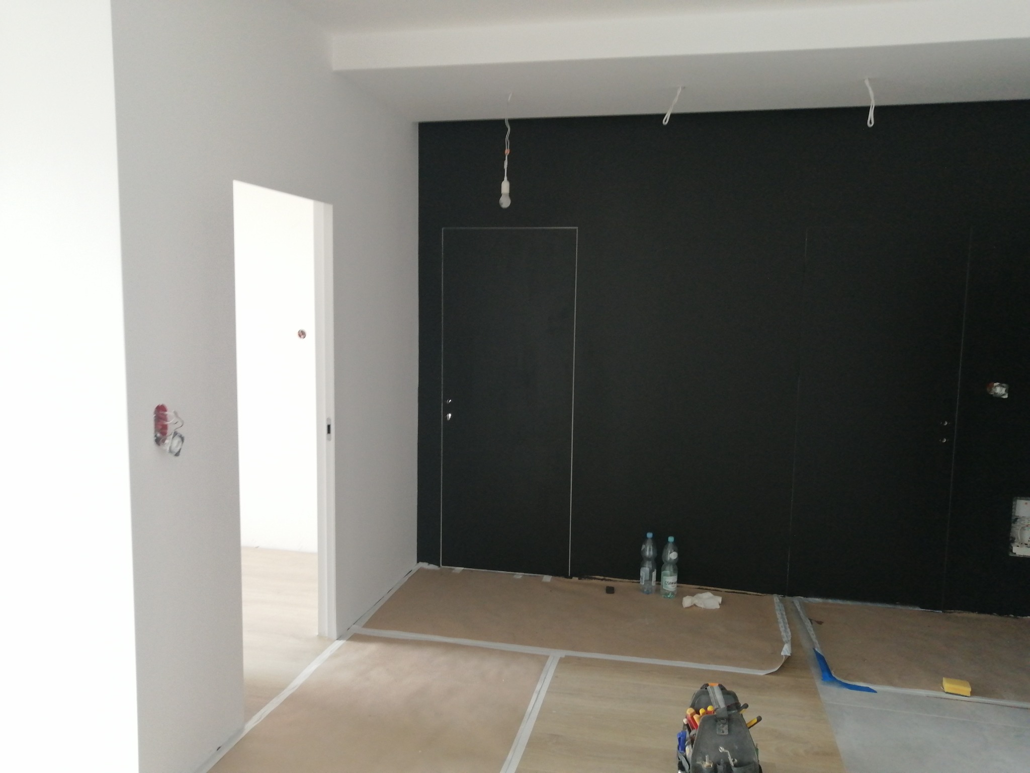 Przygotowanie do malowania ścian na ciemny kolor, zabezpieczone podłogi kartonem i taśmą, widoczne dwie butelki wody mineralnej i narzędzia w kuferku.