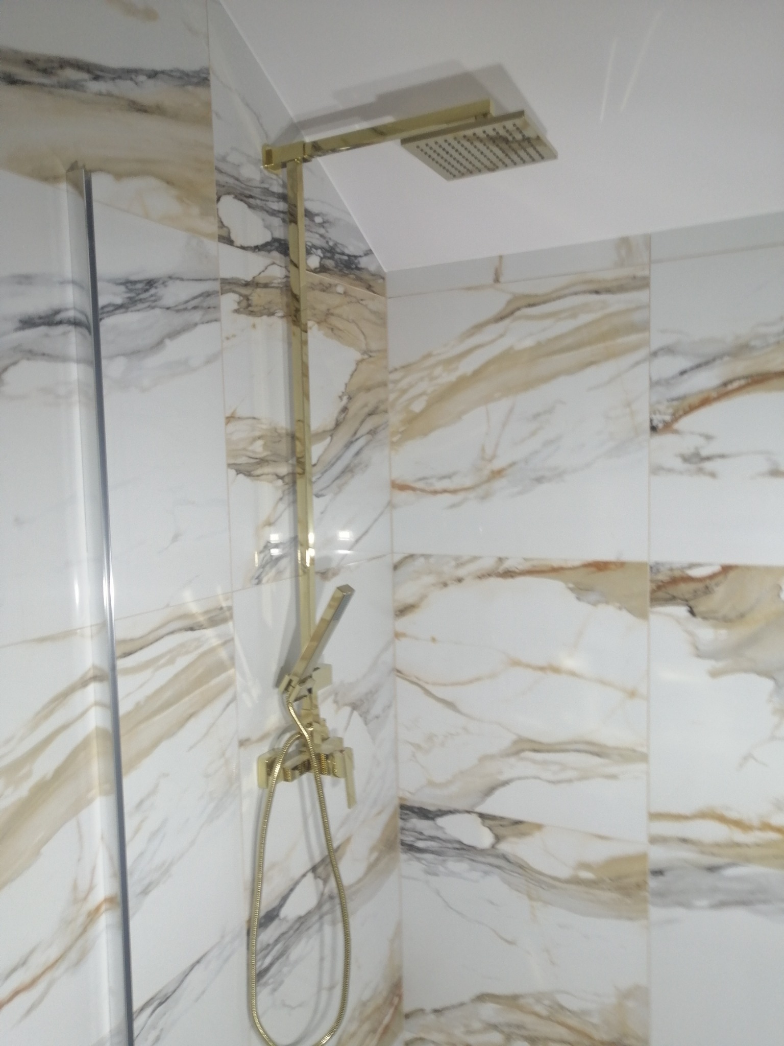 Błyszcząca, złota armatura prysznicowa z deszczownicą na ścianie wyłożonej białymi płytkami imitującymi marmur z brązowymi żyłami.