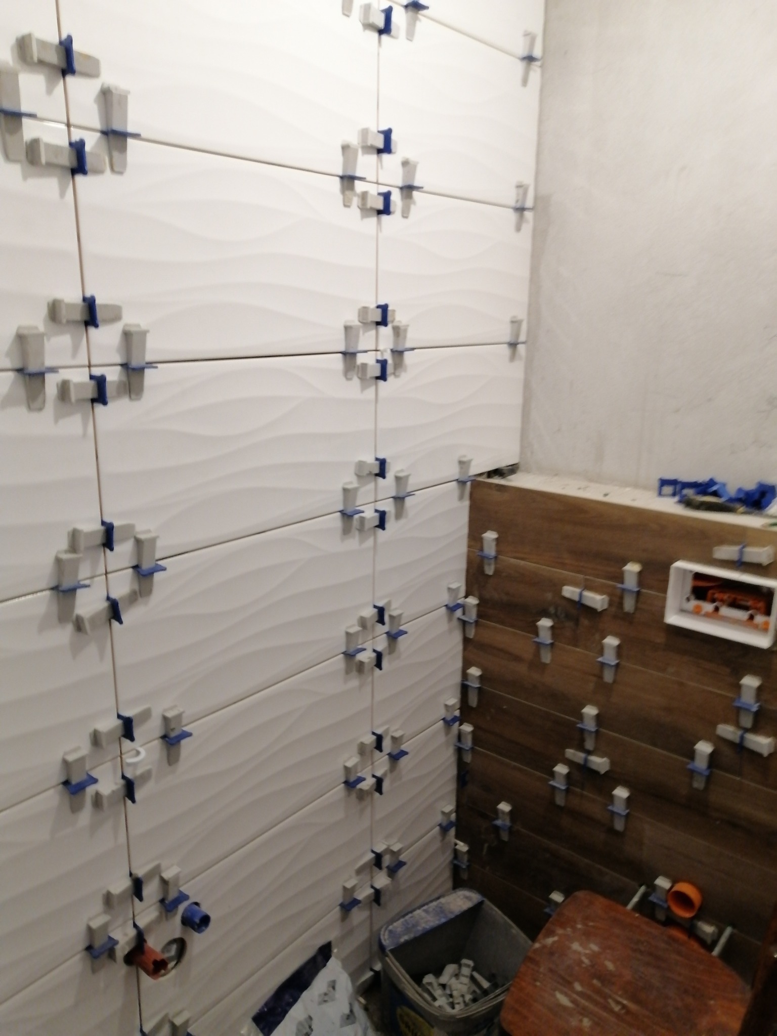 Białe, faliste płytki ścienne zamocowane krzyżykami dystansowymi w trakcie montażu, z widocznymi instalacjami hydraulicznymi i fragmentem drewnopodobnej okładziny na sąsiedniej ścianie.