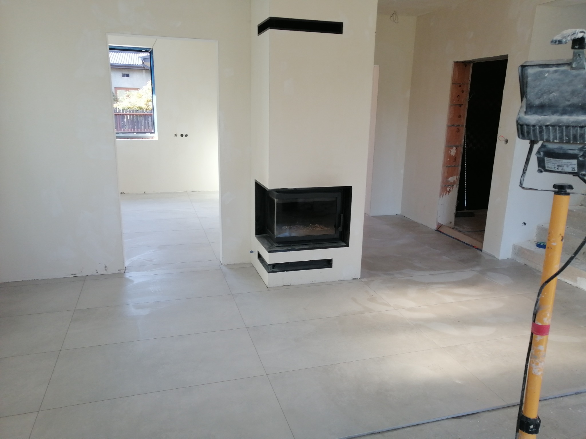 Wnętrze w trakcie wykończenia: jasne, duże płytki podłogowe, obudowa kominka z czarnymi elementami, ściany po gładzi, widoczna lampa budowlana na statywie.