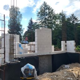 WS BUDOWNICTWO Sp. z o.o. - Wznoszenie ścian z bloczków betonu komórkowego na fundamencie z izolacją pionową, zbrojenie pionowe w narożnikach, widoczne rusztowanie i maszyna do cięcia bloczków.