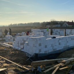 WS BUDOWNICTWO Sp. z o.o. - Wznoszenie ścian parteru domu z bloczków betonu komórkowego Ytong na fundamencie, widoczni robotnicy, teren budowy z drewnianymi odpadami, w tle zabudowa jednorodzinna i pole w porannym słońcu.