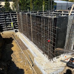 WS BUDOWNICTWO Sp. z o.o. - Zbrojenie i szalunek fundamentów pod mury oporowe, widok z góry. Elementy konstrukcyjne w wykopie, przygotowane do zalania betonem. Na pierwszym planie błoto i narzędzia.