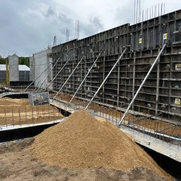 WS BUDOWNICTWO Sp. z o.o. - Szare szalunki z metalowymi podporami, świeży beton i piasek. Widok na budowę muru oporowego w pochmurny dzień. Konstrukcja zbrojona, przygotowana do zalania betonem.