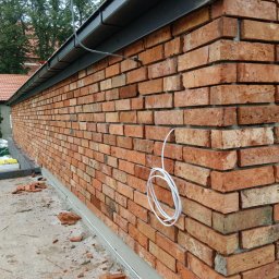 WS BUDOWNICTWO Sp. z o.o. - Ściana z czerwonej cegły klinkierowej pod metalowym dachem, z widocznym przewodem elektrycznym i drobnymi fragmentami cegieł na posadzce.