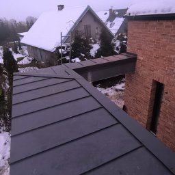 WS BUDOWNICTWO Sp. z o.o. - Nowy, grafitowy dach z blachy na domu z cegły, połączony stalową konstrukcją. W tle ośnieżone budynki i drzewa. Widok z góry, pochmurny dzień.