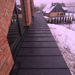 WS BUDOWNICTWO Sp. z o.o. - Nowoczesny dom z dachem z blachy na rąbek stojący, widok z góry na dach i komin z cegły klinkierowej, w tle zasypany śniegiem ogród i drugi budynek.