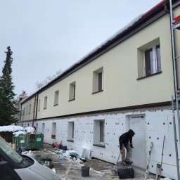 WS BUDOWNICTWO Sp. z o.o. - Ocieplanie budynku styropianem: pracownik na rusztowaniu przykłada arkusz do ściany. Widoczne okna, ośnieżone otoczenie i fragment samochodu.
