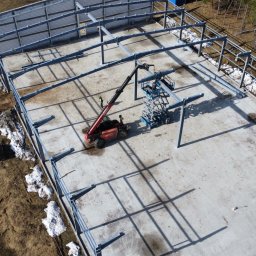 WS BUDOWNICTWO Sp. z o.o. - Montaż stalowej konstrukcji hali z użyciem podnośnika teleskopowego na betonowej płycie fundamentowej, widok z góry, częściowo ośnieżony teren.