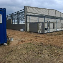 WS BUDOWNICTWO Sp. z o.o. - Budowa hali: stalowa konstrukcja, ściany z bloczków i szare panele. Niebieski kontener budowlany z oknem z lewej strony. Teren budowy, pochmurne niebo.