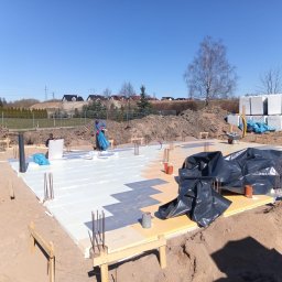 WS BUDOWNICTWO Sp. z o.o. - Fundamenty domu jednorodzinnego w trakcie izolacji termicznej, widoczne zbrojenia i przygotowane bloczki. Prace ziemne w toku, słoneczny dzień.