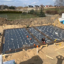 WS BUDOWNICTWO Sp. z o.o. - Zbrojenie fundamentów pod budowę domu jednorodzinnego. Widoczne stalowe pręty, folia izolacyjna i białe podkładki dystansowe. W tle zabudowa domów i drzewa.