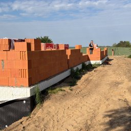 WS BUDOWNICTWO Sp. z o.o. - Wznoszenie ścian z czerwonej cegły Porotherm na fundamencie z izolacją, widoczny pracownik w tle, teren budowy z piaskiem i ogrodzeniem. Budowa domu w toku.