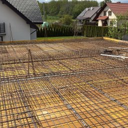 WS BUDOWNICTWO Sp. z o.o. - Zbrojenie płyty fundamentowej na żółtym szalunku, widok z góry. W tle domy jednorodzinne i zieleń. Etap budowy domu z widocznymi narzędziami.