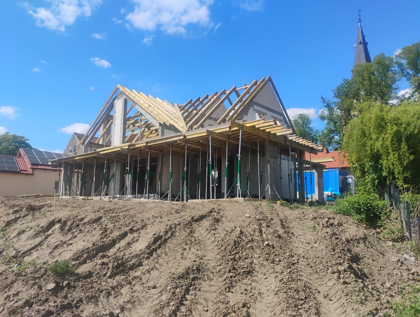 Budowa domu jednorodzinnego w trakcie realizacji: ściany z betonu komórkowego, drewniana więźba dachowa, podpory szalunkowe. W tle wieża kościoła i błękitne niebo.