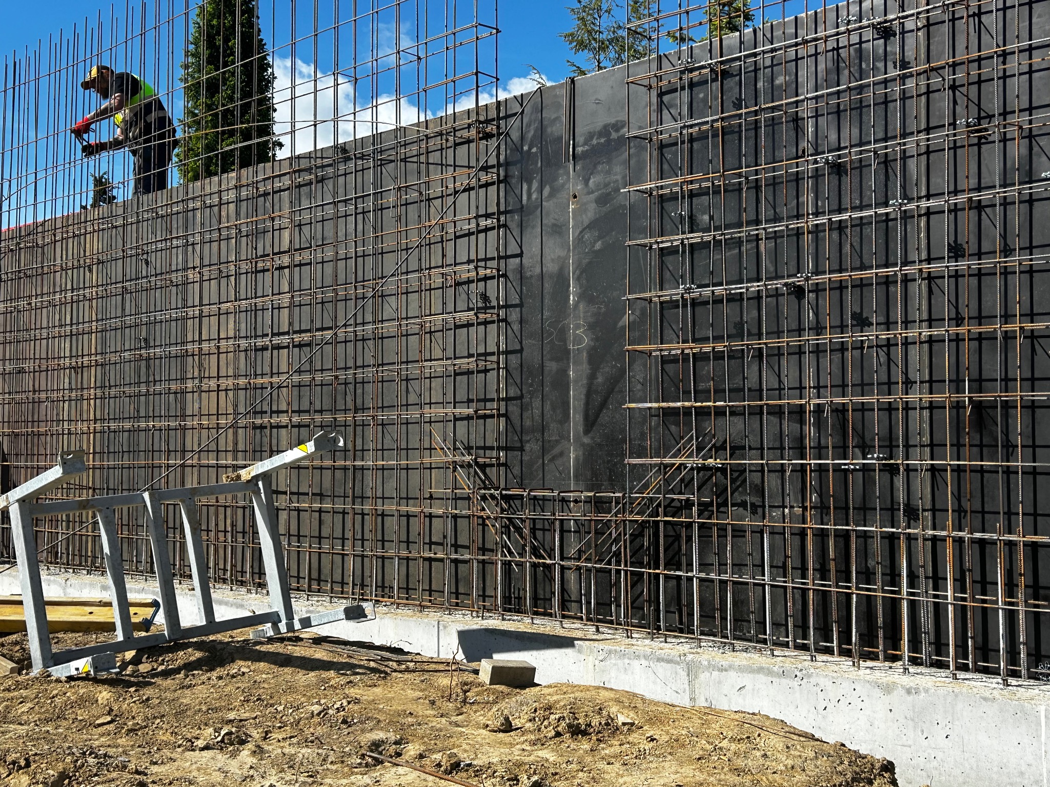 Zbrojenie ściany oporowej z widocznym pracownikiem na tle błękitnego nieba. Na pierwszym planie leżąca drabina. Detal budowy w trakcie realizacji.