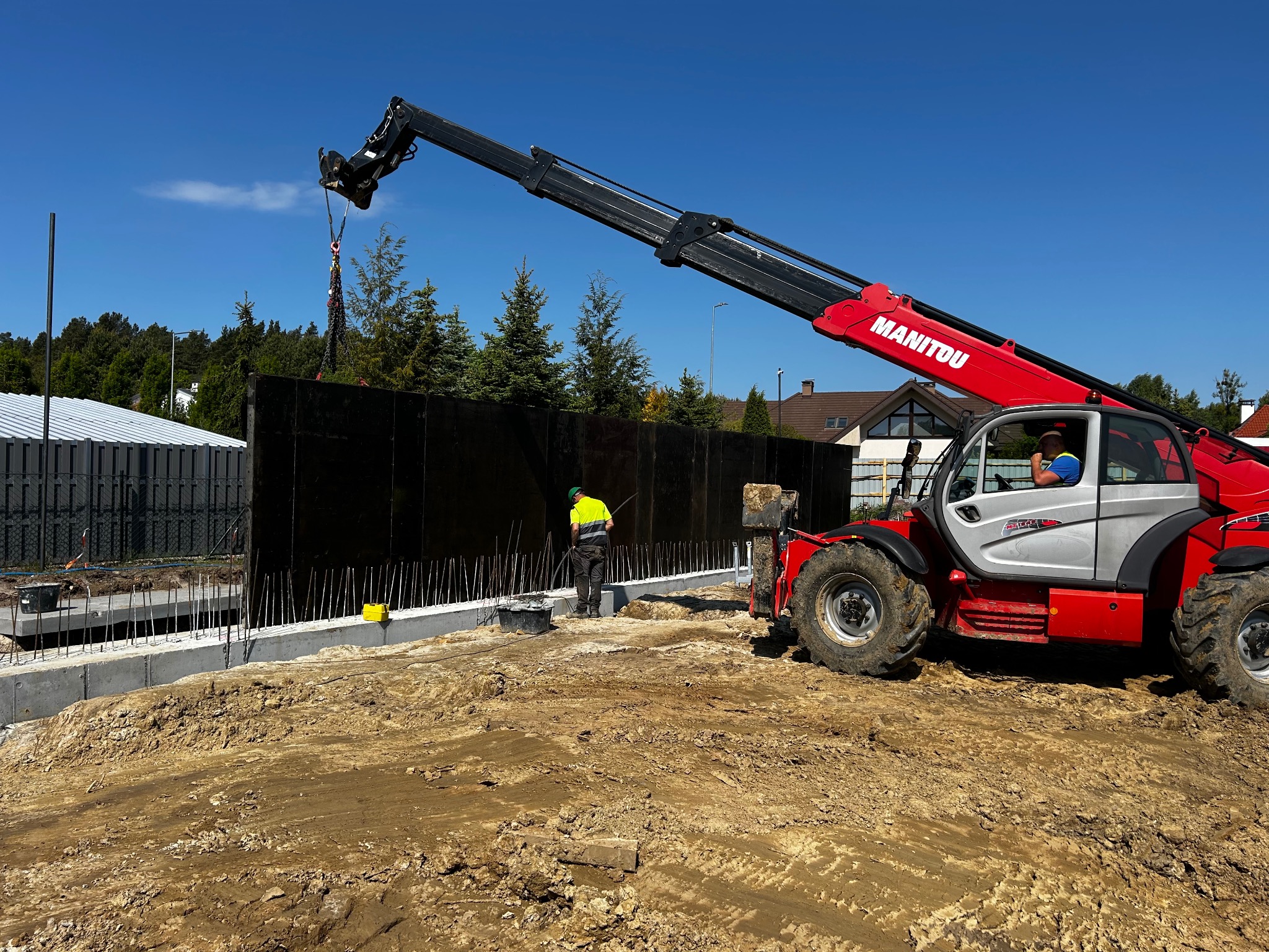 Montaż paneli szalunkowych na fundamencie z użyciem podnośnika teleskopowego Manitou, pracownik w odblaskowej kamizelce nadzoruje proces.