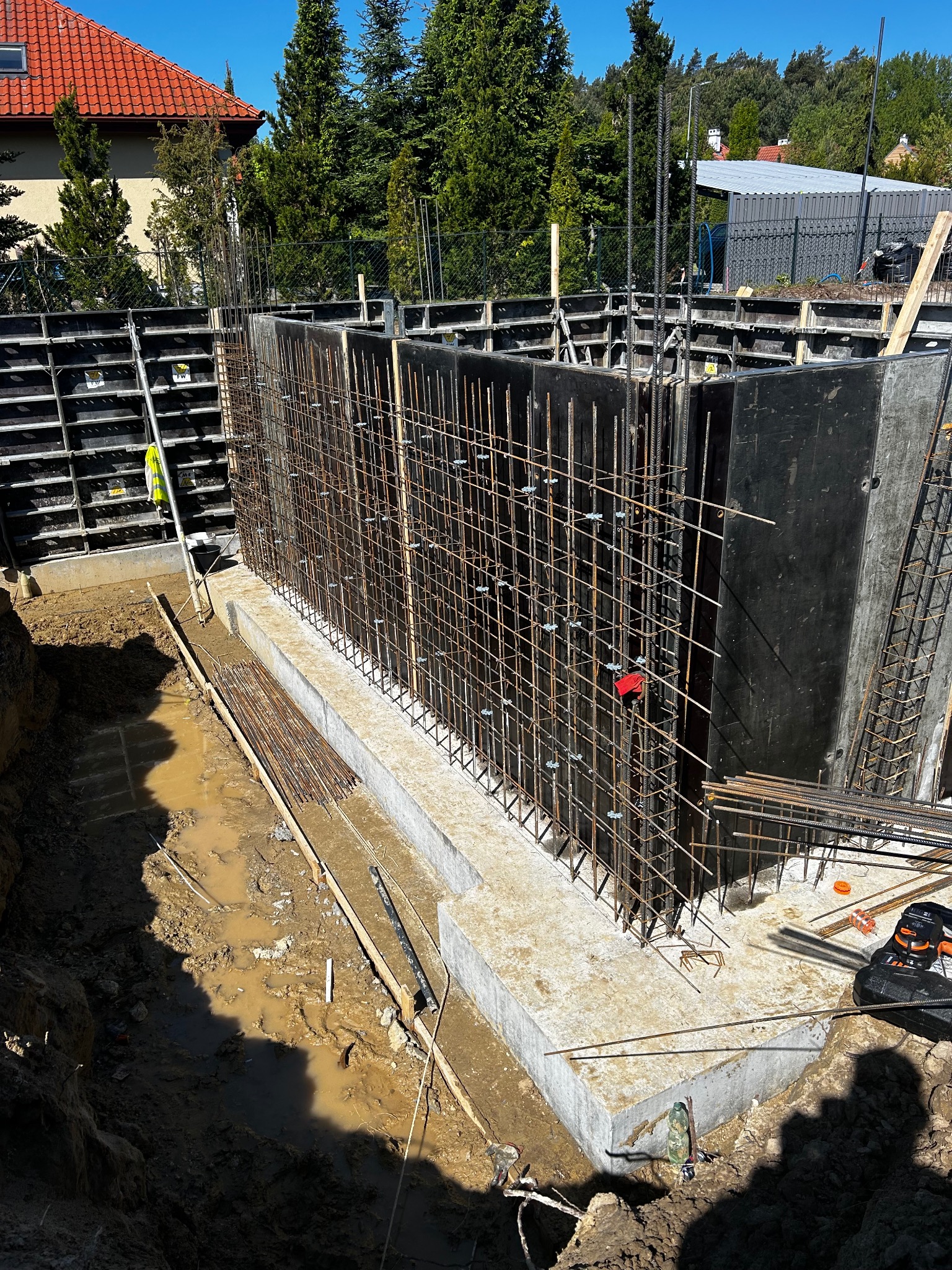 Zbrojenie i szalunek fundamentów pod mury oporowe, widok z góry. Elementy konstrukcyjne w wykopie, przygotowane do zalania betonem. Na pierwszym planie błoto i narzędzia.