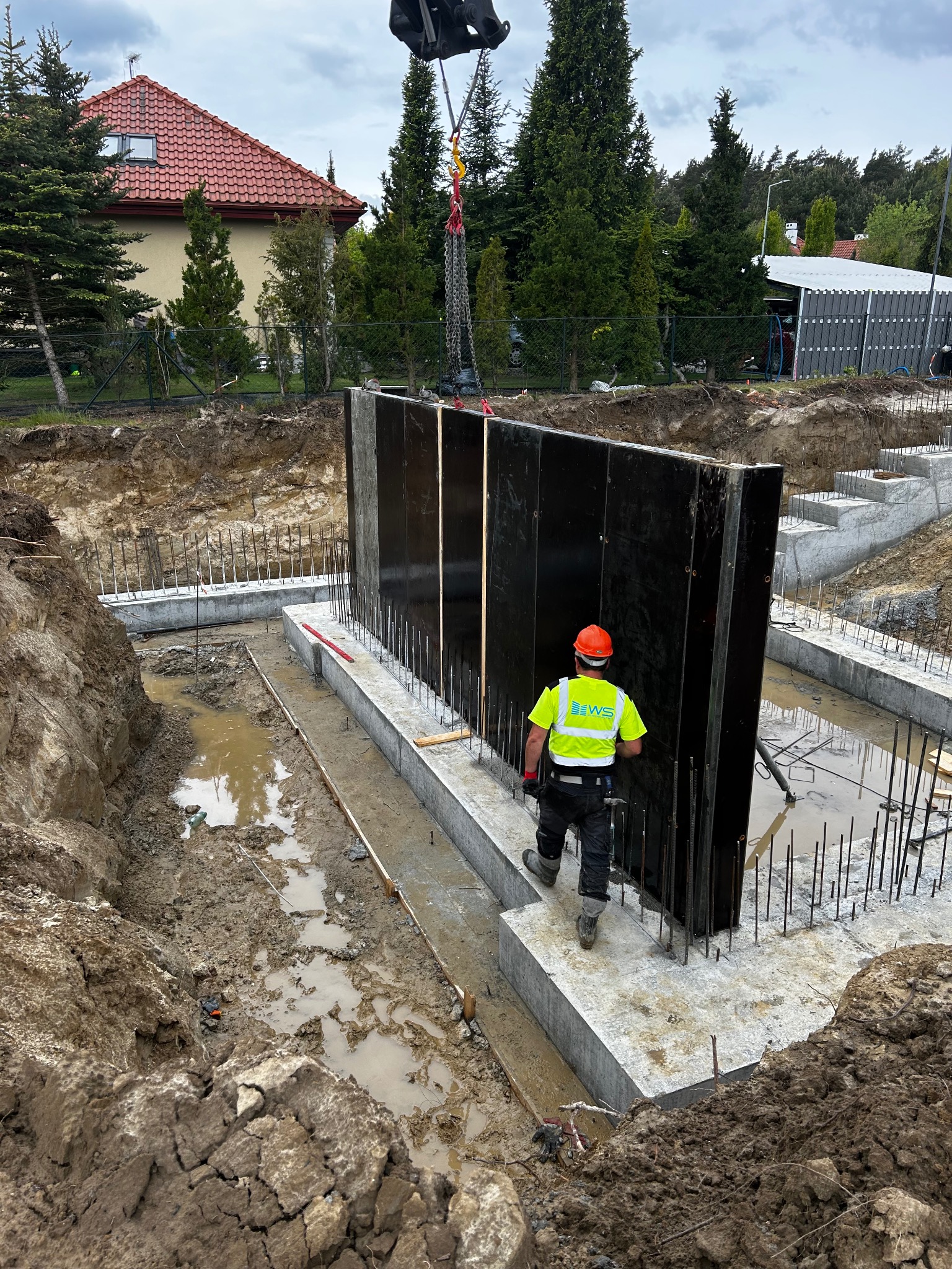 Montaż szalunków na fundamencie domu jednorodzinnego. Pracownik w kasku i kamizelce odblaskowej nadzoruje proces. Widoczne zbrojenia i wykop pod budynek.