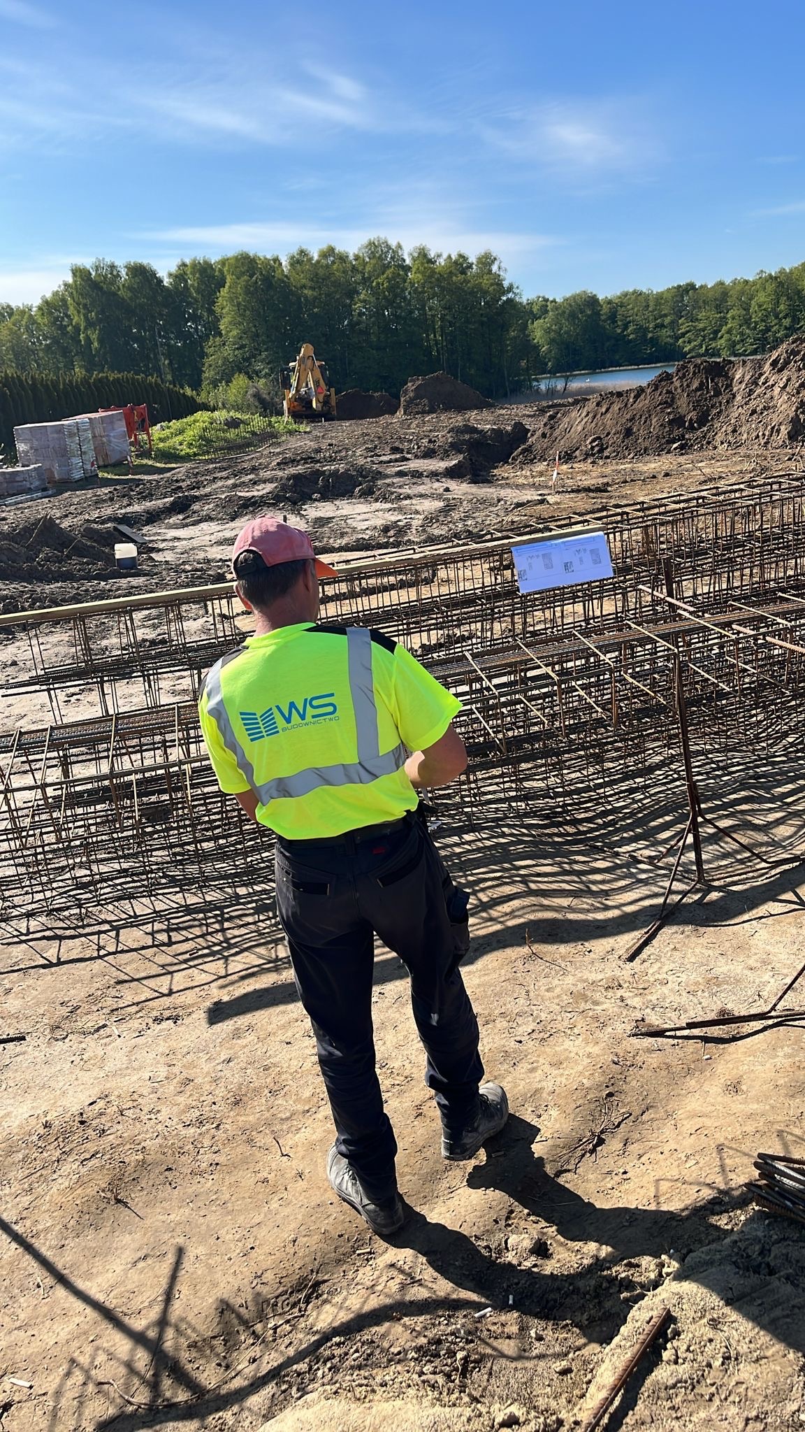 Pracownik w żółtej kamizelce z logo WS Budownictwo analizuje plan na tablecie, stojąc przed zbrojeniem fundamentów na placu budowy z koparką w tle.