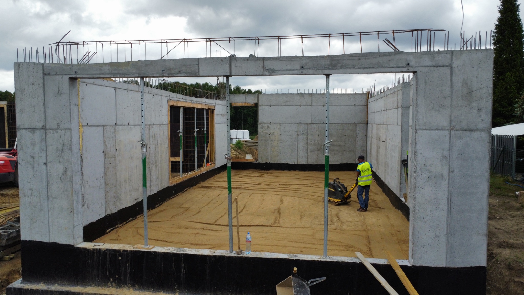 Wznoszenie ścian domu z prefabrykatów betonowych. Pracownik zagęszcza podłoże zagęszczarką. Widoczne zbrojenia i podpory. Surowy stan.